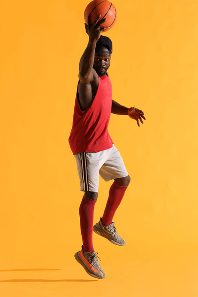 Full length portrait of black man in red shirt, black cap and grey shorts playing basketball. Studio, yellow background - Foto, Bild