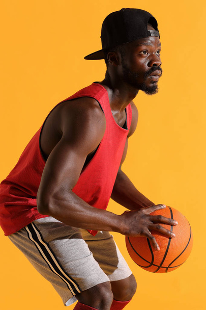 Man in red shirt and black cap hold basketball ball in hands in front of him. Studio, yellow background - Zdjęcie, obraz
