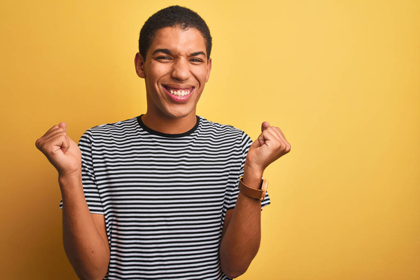 Joven hombre árabe guapo con camiseta a rayas de la marina sobre fondo amarillo aislado celebrando sorprendido y sorprendido por el éxito con los brazos levantados y los ojos abiertos. Concepto ganador
. - Foto, imagen
