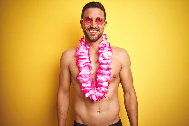 Genç yakışıklı gömleksiz adam yüzünde mutlu ve serin bir gülümseme ile sarı arka plan üzerinde güneş gözlüğü ve pembe hawaiian lei giyiyor. Şanslı kişi. - Fotoğraf, Görsel