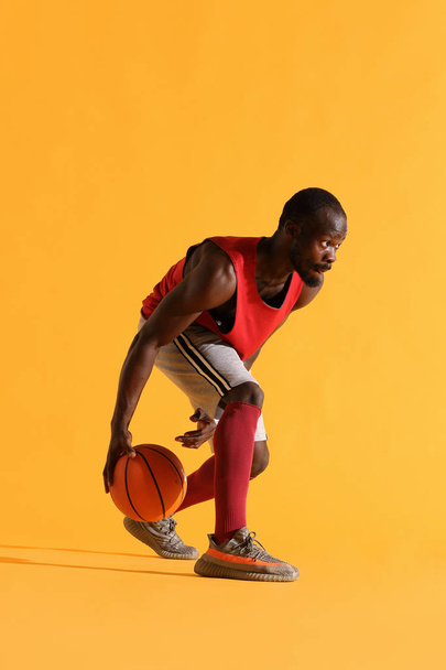 Full length portrait of black man in red shirt, black cap and grey shorts playing basketball. Studio, yellow background - Photo, Image