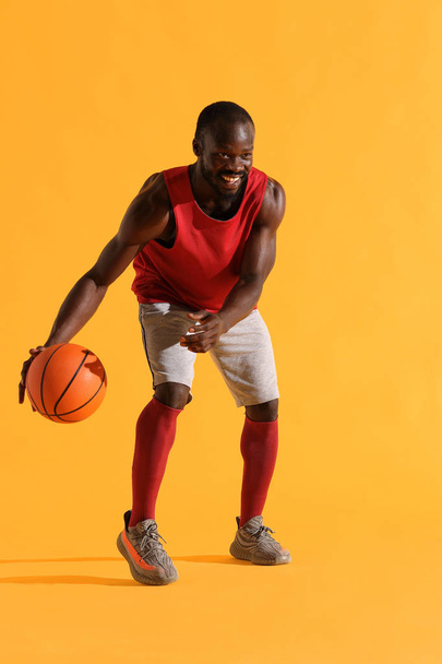 Full length portrait of black man in red shirt, black cap and grey shorts playing basketball. Studio, yellow background - Foto, Bild