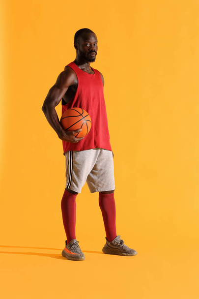 Full length portrait of black man in red shirt, black cap and grey shorts playing basketball. Studio, yellow background - Фото, изображение