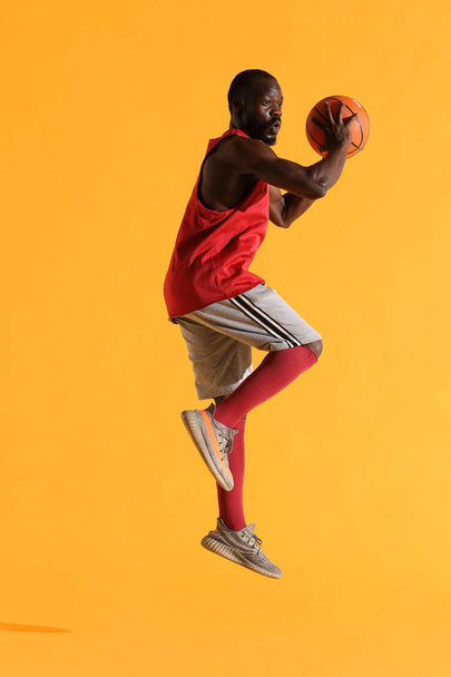 Full length portrait of black man in red shirt, black cap and grey shorts playing basketball. Studio, yellow background - Fotoğraf, Görsel
