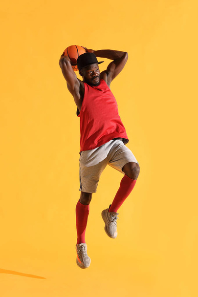 Full length portrait of black man in red shirt, black cap and grey shorts playing basketball. Studio, yellow background - Foto, Imagen