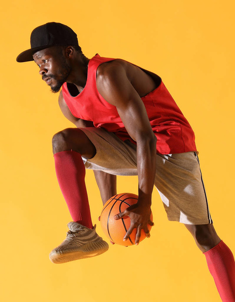 Sport portrait of black man in red shirt and grey shorts playing basketball. Studio, yellow background - Фото, зображення