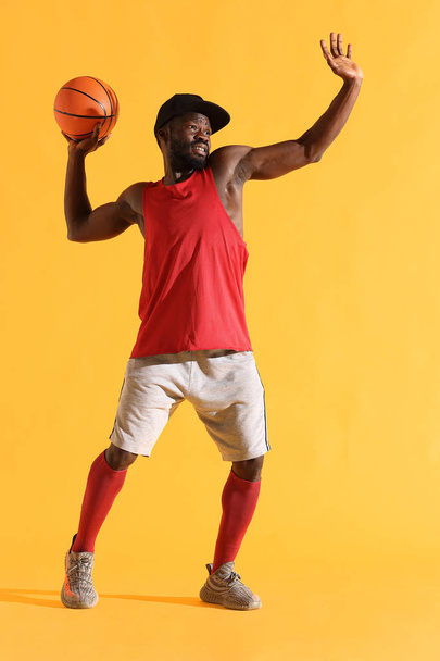 Full length portrait of black man in red shirt, black cap and grey shorts playing basketball. Studio, yellow background - Zdjęcie, obraz