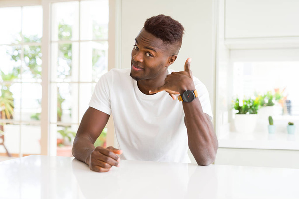 自宅の白いテーブルの上のハンサムなアフリカ系アメリカ人男性は、電話で話すような手と指で電話ジェスチャーをして微笑んでいます。概念の伝達. - 写真・画像