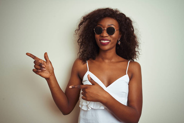 Jonge Afro-Amerikaanse vrouw draagt t-shirt en zonnebril over geïsoleerde witte achtergrond glimlachend en kijkend naar de camera die met twee handen en vingers naar de zijkant wijst. - Foto, afbeelding