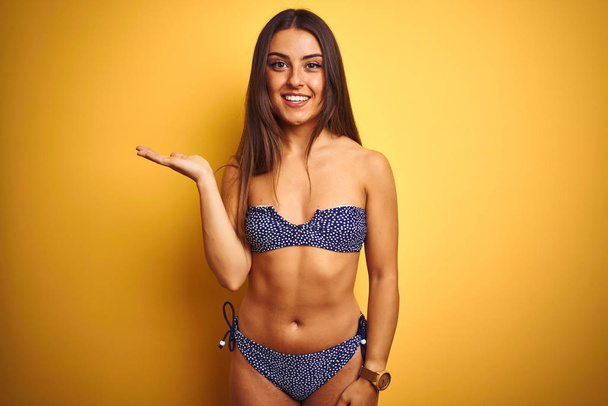 Young beautiful woman on vacation wearing bikini standing over isolated yellow background smiling cheerful presenting and pointing with palm of hand looking at the camera. - Photo, Image