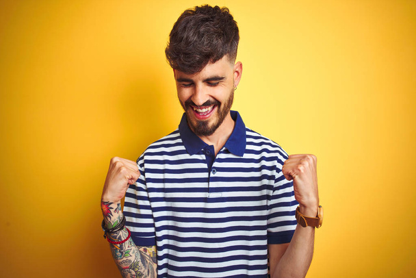 Young man with tattoo wearing striped polo standing over isolated yellow background very happy and excited doing winner gesture with arms raised, smiling and screaming for success. Celebration concept. - Photo, Image