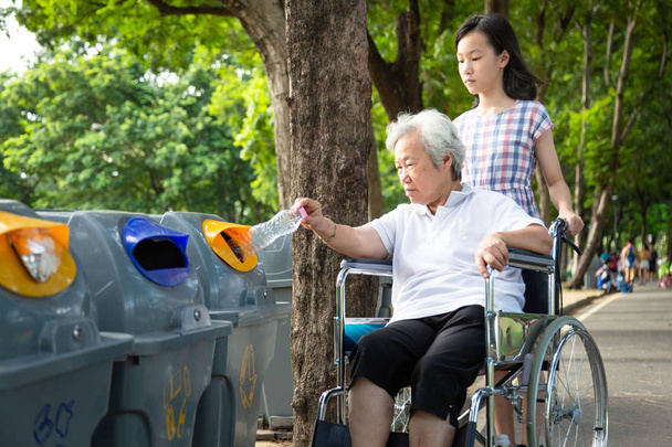 Asyalı küçük kız ya da torun ve yaşlı büyükanne geri dönüşüm kutusuna plastik su şişesi koyuyorlar, yaşlı turist tekerlekli sandalyede, çöp kutusuna çöp atıyorlar, çevre koruma, küresel ısınma kavramı.  - Fotoğraf, Görsel