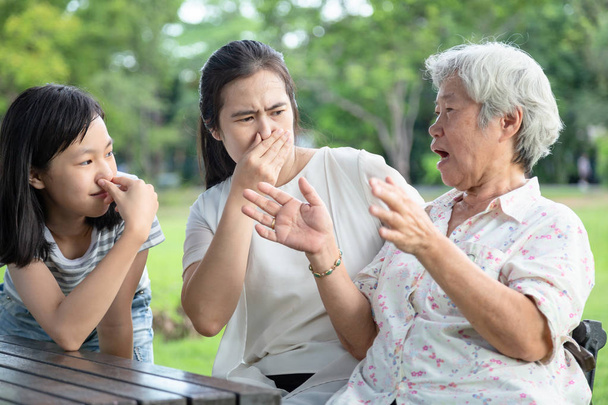 asiatische Großmutter, die mit ihrer Familie spricht, älteren Menschen Mundgeruch, Tochter, Enkelin, die ihre Nase schließt, sehr schlechter Geruch, Frau und Kind spüren Gestank, Gesichtsausdruck, Zahngesundheit, Mundgeruch-Konzept - Foto, Bild
