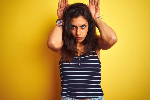 Young beautiful woman wearing striped t-shirt standing over isolated yellow background Doing bunny ears gesture with hands palms looking cynical and skeptical. Easter rabbit concept. - Photo, Image