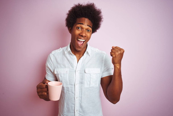 Giovane uomo afroamericano bere una tazza di caffè in piedi sopra isolato sfondo rosa urlando orgoglioso e celebrando la vittoria e il successo molto eccitato, emozione tifo
 - Foto, immagini