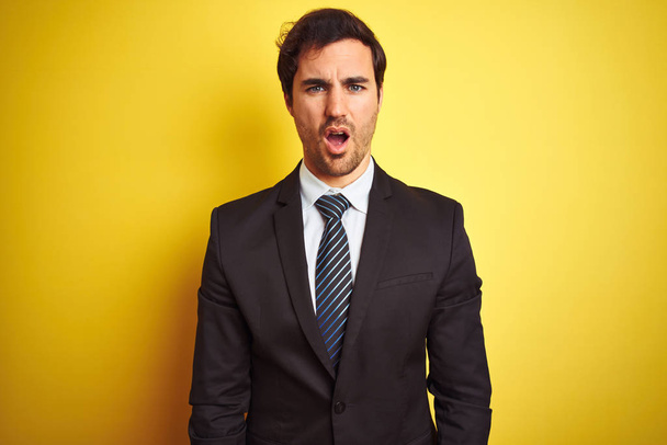Young handsome businessman wearing suit and tie standing over isolated yellow background In shock face, looking skeptical and sarcastic, surprised with open mouth - Fotografie, Obrázek
