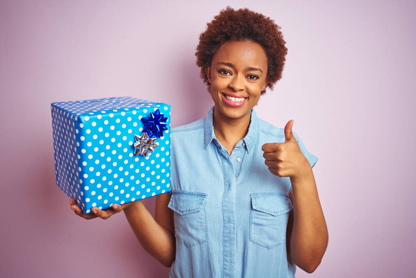 African American kobieta trzyma prezent urodzinowy nad różowym białym tle szczęśliwy z wielkim uśmiechem robi OK znak, kciuk w górę z palcami, doskonały znak - Zdjęcie, obraz