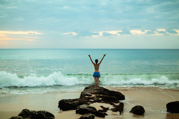 aufgeregte junge Frau, die am Strand vor dem Ozean die Arme hebt. Blick von hinten. Sonnenuntergang am Strand. bali, indonesien. - Foto, Bild