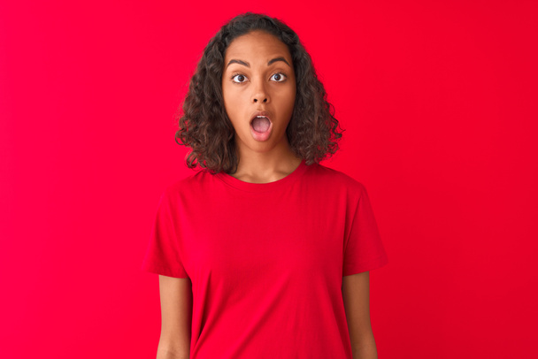Νέα βραζιλιάνικη γυναίκα φορώντας t-shirt στέκεται πάνω από απομονωμένο κόκκινο φόντο φοβισμένος σε σοκ με μια έκπληξη πρόσωπο, φοβισμένος και ενθουσιασμένος με την έκφραση του φόβου - Φωτογραφία, εικόνα