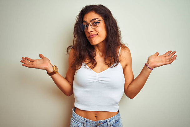 Young beautiful woman wearing t-shirt and glasses standing over isolated white background clueless and confused expression with arms and hands raised. Doubt concept. - Photo, image