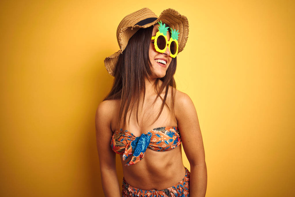 Nainen lomalla yllään bikinit ja ananas aurinkolasit yli eristetty keltainen tausta etsii sivuun hymyillen kasvot, luonnollinen ilme. Nauraen itsevarmana
. - Valokuva, kuva