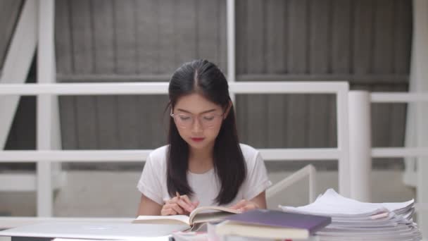 アジアの学生の女性は大学の図書館で本を読みます。大学キャンパス構想の講義デスクで知識を学びながら疲れた若い学部生の女の子が問題を抱えている. - 映像、動画