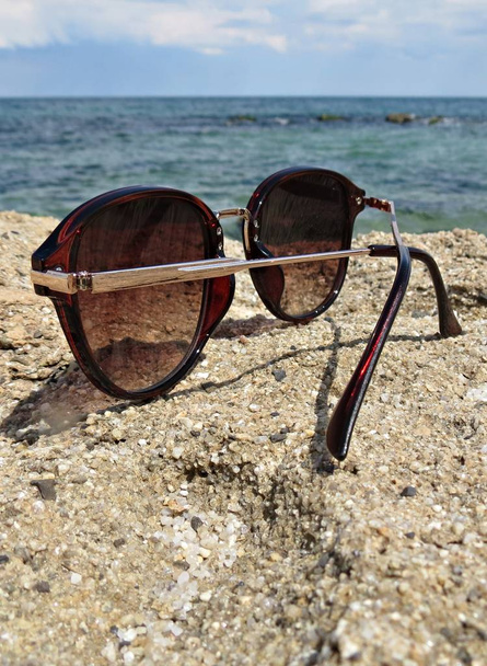 Крупним планом жіночі сонцезахисні окуляри на скелі біля моря. Коричневі сонцезахисні окуляри, розміщені на скелях і морському фоні
 - Фото, зображення