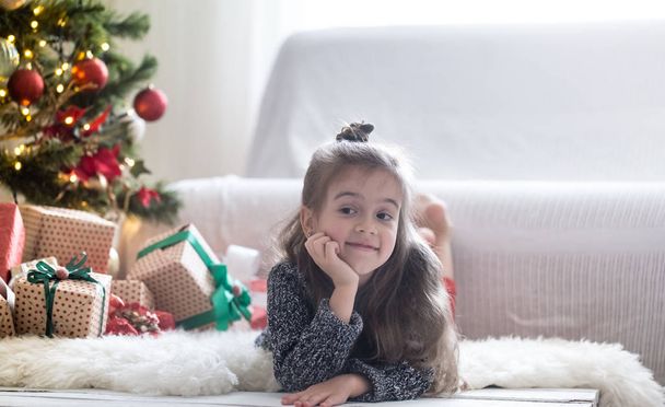 hübsches kleines Mädchen liegt auf dem Boden in ihrem Zimmer zu Hause in der Nähe des Weihnachtsbaums - Foto, Bild