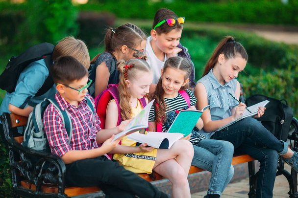 Happy Schoolmates muotokuva. Koulukaverit istuu kirjoja puinen penkki kaupungin puistossa ja opiskelee aurinkoisena päivänä. - Valokuva, kuva