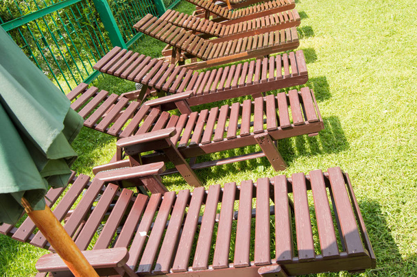 Wooden deck chairs stand in a row on the green grass under the sun in the recreation area on the private territory of a modern luxury hotel. Garden for sunbathing and relaxation - Photo, Image
