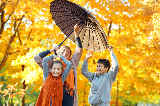 傘の下でポーズをとる幸せな家族は、秋の都市公園で遊んで楽しんでいます。子供と親が一緒に素敵な一日を過ごします。木々の明るい日差しと黄色い葉、秋の季節. - 写真・画像