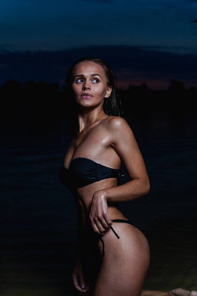 Beautiful model in the water. In a black bikini swimsuit for the night, swimming and posing. - Photo, Image
