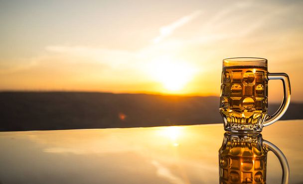 Szklanka piwa na plaży o zachodzie słońca. Koncepcja letniego napoju chłodzącego. Zbliżenie szklankę piwa zanurzenie z bokeh Sunlight tle - Zdjęcie, obraz