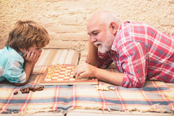 Ребенок играет в шахматы. Дедушка и внук играют в шахматы и улыбаются, проводя время вместе дома. Шахматная конкуренция
. - Фото, изображение