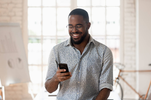 Happy black businessman holding smartphone using apps standing in office - Photo, Image