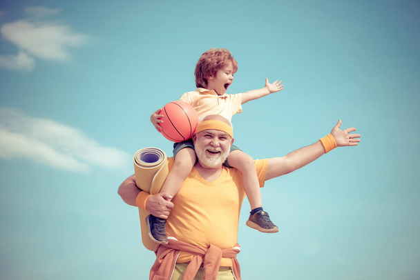 Senior man and cute little boy exercising on blue sky background - isolated. Father and son sporting - family time together. Doing sports is free. - 写真・画像