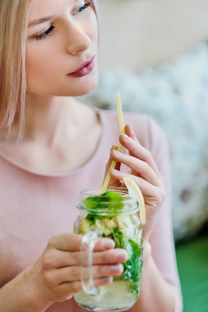 Joven chica rubia hermosa en un vestido rosa se sienta en una mesa de café, bebe limonada de un frasco de vidrio
. - Foto, Imagen