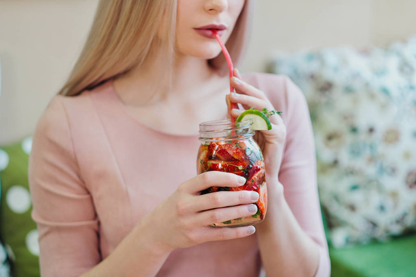 Mladá krásná blondýnka v růžových šatech sedí u stolku v kavárně, pije limonádu ze skleněného džbánu. - Fotografie, Obrázek