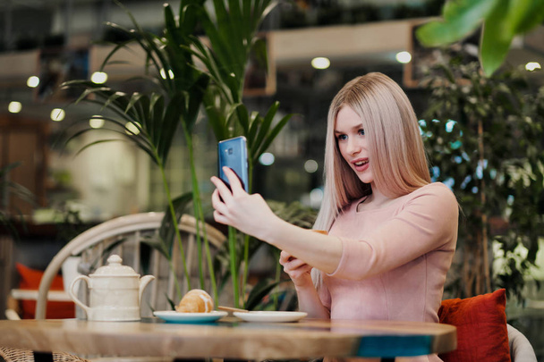 Молодая красивая девушка сидит в зеленом кафе за столом. Пить чай с круассанами, болтать, смеяться и фотографировать на смартфон
. - Фото, изображение