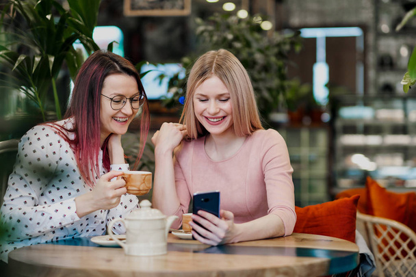 Two young beautiful girls sit in a green cafe at a table. Drink tea with croissants, chatting, laughing and taking pictures on a smartphone. - Foto, afbeelding