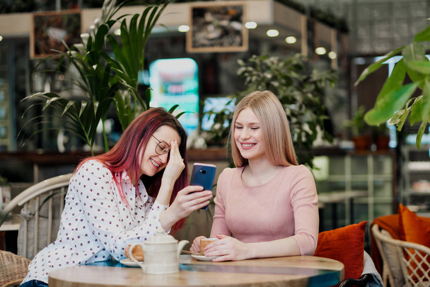 Dos chicas hermosas jóvenes se sientan en un café verde en una mesa. Beba té con croissants, charlando, riendo y tomando fotos en un teléfono inteligente
. - Foto, Imagen