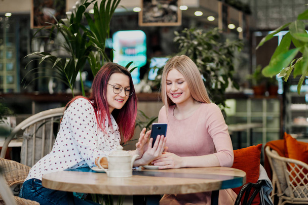 Two young beautiful girls sit in a green cafe at a table. Drink tea with croissants, chatting, laughing and taking pictures on a smartphone. - Foto, Imagem
