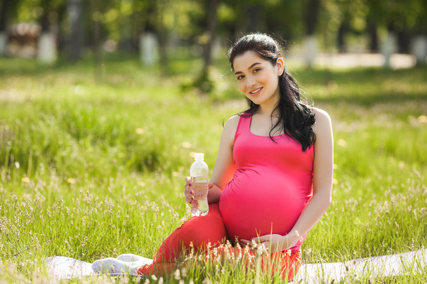 Young pregnant woman doing fitness exercises outdoors and drinking clean bottled water. Expecting female doing yoga on the fresh air. - Photo, Image
