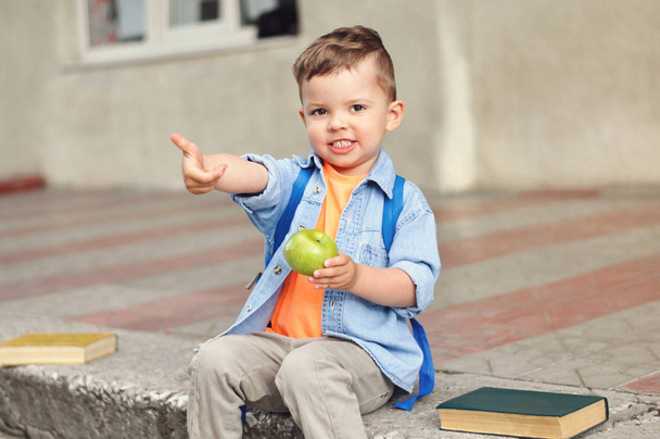 Mały trzyletni przedszkolaka z plecakiem na plecach siedzi w pobliżu szkoły i pokazuje kciuka w górę  - Zdjęcie, obraz