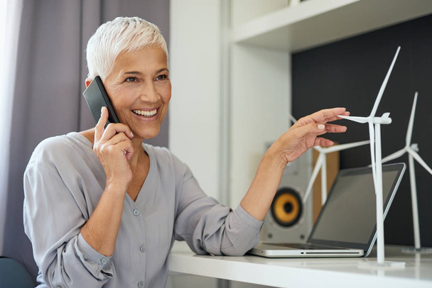 Gorgeous Caucasian senior woman talking on the phone and touching windmill model while sitting in office. Start up business concept. - Foto, Imagem