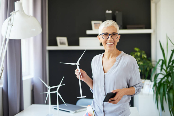Beautiful smiling Caucasian senior woman standing in office, holding windmill model and smart phone. Start up business concept. - Zdjęcie, obraz