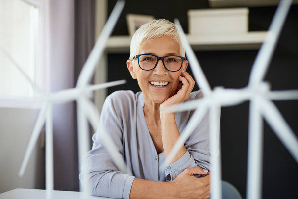 Happy Caucasian senior woman leaning on desk and looking at camera. On desk are windmill models. Start up business concept. - Фото, зображення