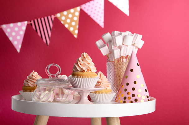 Table with party hat, cupcake and other sweets on burgundy background. Candy bar - 写真・画像