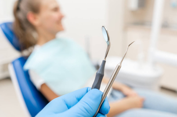Close-up hand of dentist in the glove holds tool probe and mirror. The patient in the dental chair at the background. Dental work in clinic. Office where dentist conducts inspection and concludes. - Φωτογραφία, εικόνα