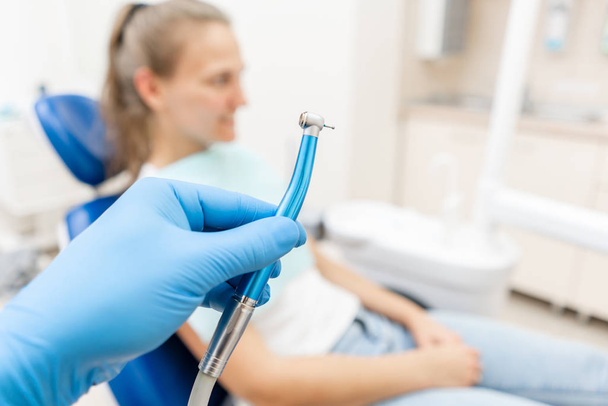 Close-up hand of dentist in the glove holds dental high speed turbine. The patient in blue chair at the background. Office where dentist conducts inspection and concludes. - Foto, Imagen